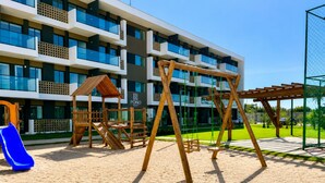 Un terrain de jeux avec des balançoires et un toboggan, une cabane en bois et, au fond, un bâtiment doté de balcons.