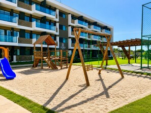 A playground with swings and a slide, a wooden playhouse, and a building with balconies in the background. - Spacious 1-bedroom apartment with AC in perfect Ipojuca (Ipojuca)