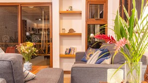 Un salon comprenant un canapé, une table basse, un vase avec des fleurs, une bibliothèque et une fenêtre donnant sur la cuisine.