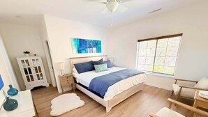Een slaapkamer met een bed, nachtkastje, stoel, raam met zonwering en een plafondventilator.