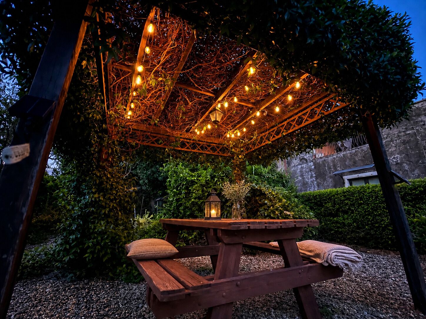 A wooden picnic table with cushions, surrounded by greenery and a decorative overhead structure with lights.