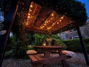 A wooden picnic table with cushions, surrounded by greenery and a decorative overhead structure with lights. - Loft Sole – A Retreat with a Pool by Sicily Escape (Sicilia)