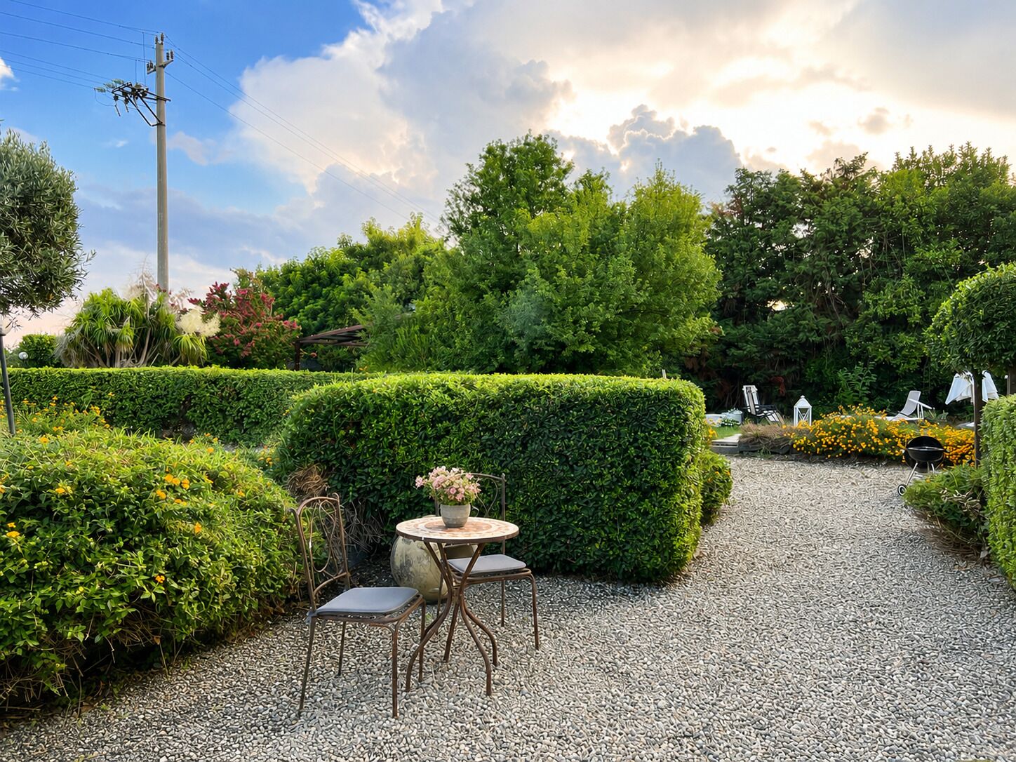 Un chemin de jardin bordé d’une table et de chaises, entouré de haies soigneusement taillées et de diverses plantes.