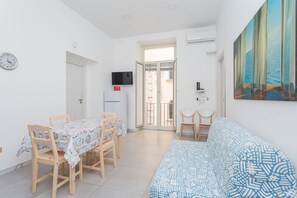 Ein Zimmer mit Esstisch, Stühlen, einer Couch, einem Fernseher und einer Tür zu einem Balkon.