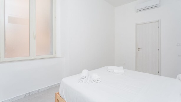 Um quarto branco com uma cama, janela, ar-condicionado e porta.