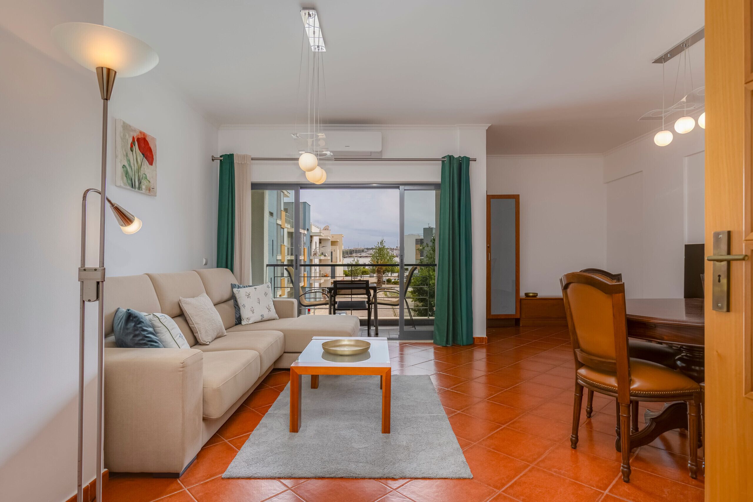 Een moderne woonkamer met een bank, salontafel, eettafel en stoelen. Er is een groot raam met gordijnen, en een balkon is zichtbaar.