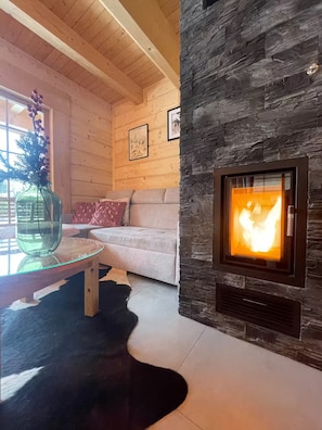 A cozy living room with a stone fireplace, wooden ceiling beams, a glass coffee table, and a sofa with cushions.