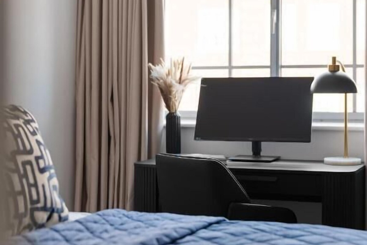 A hotel room with a bed, a desk with a computer, a lamp, and a window with curtains.
