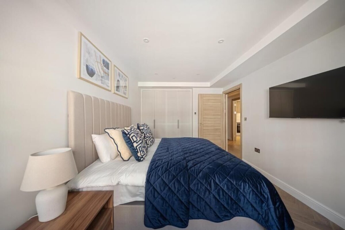 A bedroom with a bed, a bedside lamp, a TV on the wall, and a door leading to another room.