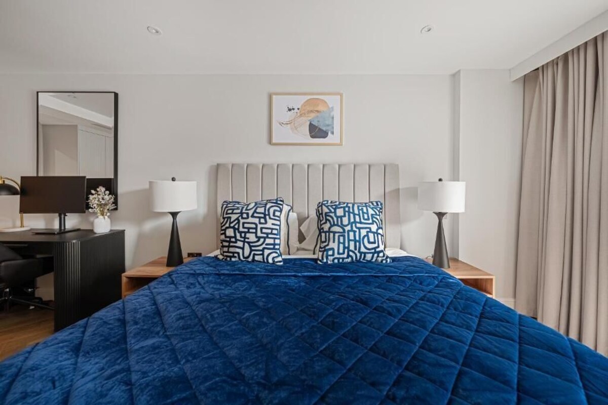A hotel room with a bed, two nightstands, a desk, a chair, a mirror, and a painting on the wall.