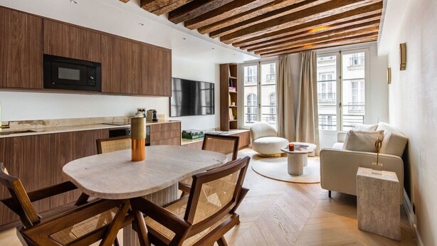 A modern kitchen with wooden beams, a dining area with a round table, and a living space with a sofa and a TV. - 1-Bedroom-Louvre-Saint-Honoré-Palais-Royal-AC (Paris)