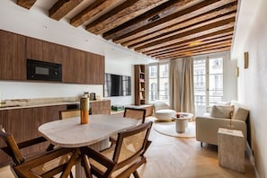A modern kitchen with wooden beams, a dining area with a round table, and a living space with a sofa and a TV. - 1-Bedroom-Louvre-Saint-Honoré-Palais-Royal-AC (Paris)