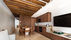 A modern kitchen with wooden cabinets, a dining area with a round table and chairs, a flat-screen TV mounted on the wall, and a white sofa in the living area. - 1-Bedroom-Louvre-Saint-Honoré-Palais-Royal-AC (Paris)