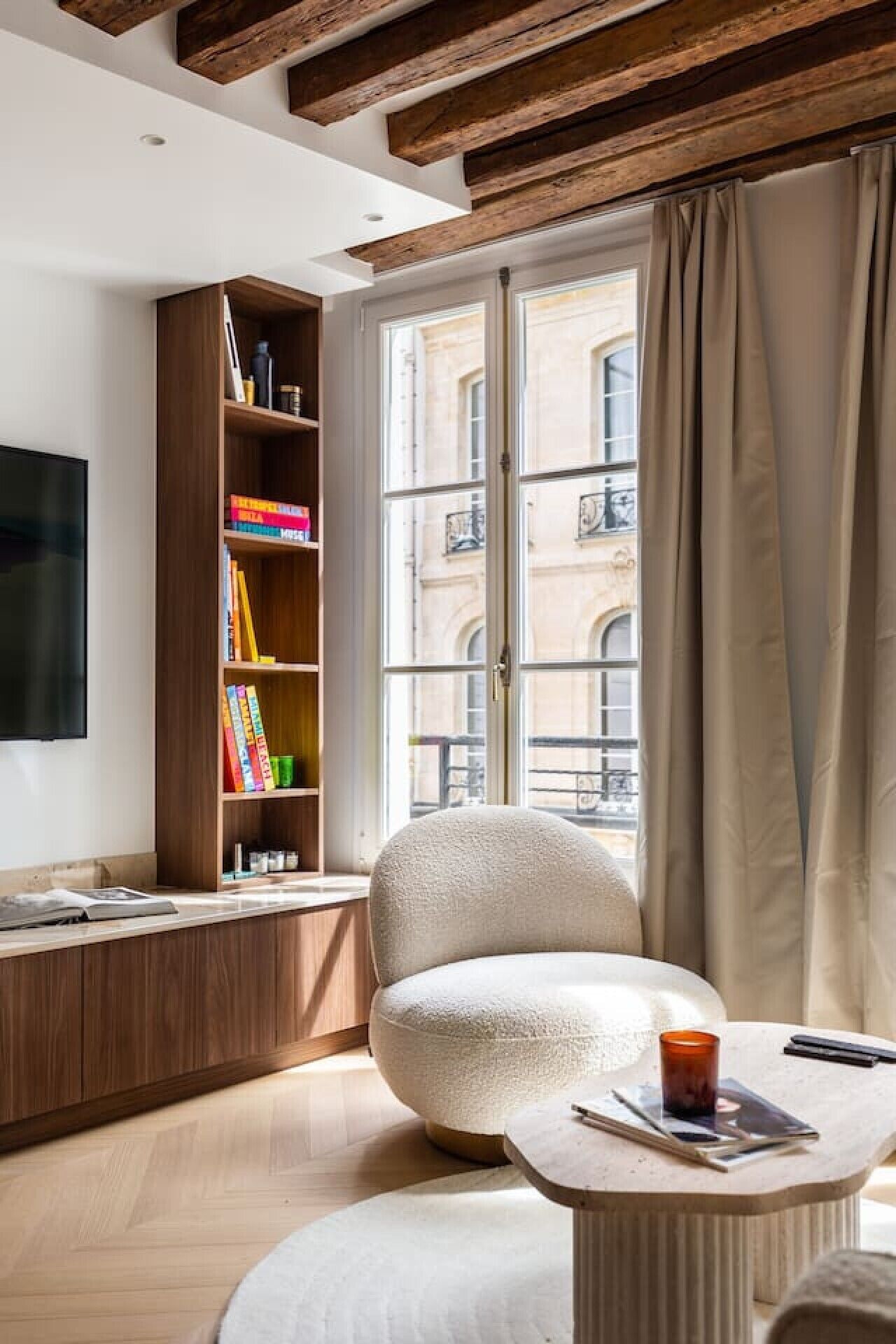 A modern living room with a wooden bookshelf, a comfortable armchair, a round coffee table, and a large window with curtains.