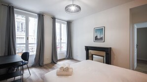Una habitación de hotel con cama, escritorio, silla, chimenea y ventanas grandes con cortinas.