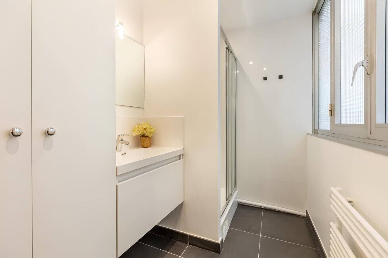 A modern bathroom with a white vanity, a glass shower enclosure, and a window with blinds.