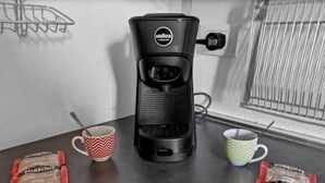 Eine Kaffeemaschine auf einem Tisch, dazu zwei Tassen und zwei Kaffeepäckchen.