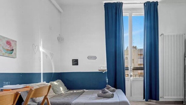 Ein Schlafzimmer mit Bett, Schreibtisch und Stuhl. Ein Fenster mit Vorhängen, eine Heizung und ein Schild an der Wand.