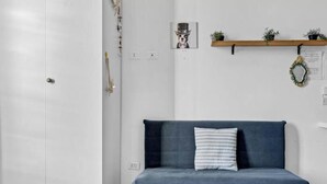 Ein Zimmer mit einem dunkelblauen Sofa, einer weißen Tür, einem Holzregal mit Pflanzen und einem gerahmten Bild an der Wand.