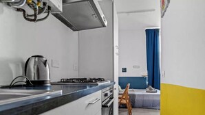 Eine kleine Küche mit Herd, Wasserkocher und Schränken, die in einen Schlafbereich mit Bett und blauem Vorhang übergeht.