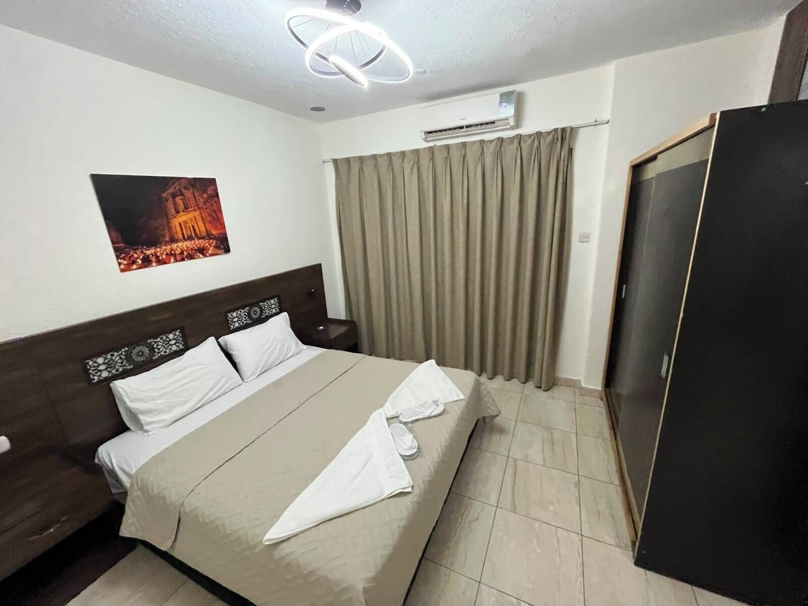 A hotel room with a bed, a nightstand, an air conditioning unit, a door, and a curtain.