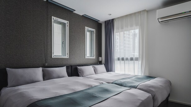 Une chambre d’hôtel avec deux lits, un mur à texture sombre, une fenêtre avec des rideaux et un appareil de climatisation.