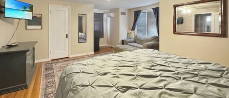 A bedroom with a bed, a ceiling fan, a TV, a dresser, a rug, and a door leading to another room.
