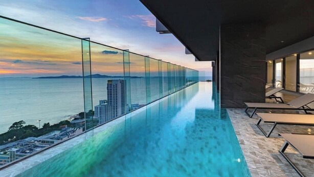 Una piscina infinita moderna con barandilla de vidrio, sillas de descanso y vista al mar y edificios a lo lejos.