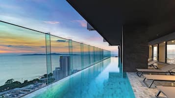 Une piscine à débordement moderne, dotée d’une paroi en verre, de chaises longues et offrant une vue sur la mer et les bâtiments lointains.