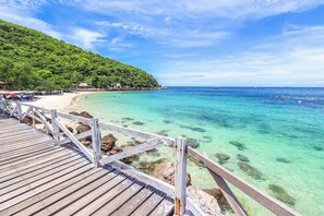 Eine Holzstege führt zu einem Strand mit klarem türkisblauem Wasser und einem bewaldeten Hügel im Hintergrund.