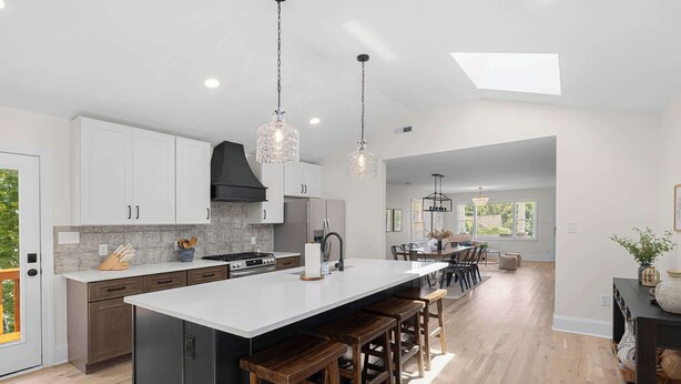 A modern kitchen with white cabinets, a large island, wooden stools, and pendant lighting.