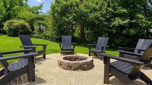 A fire pit surrounded by Adirondack chairs in a garden with trees and a lawn.