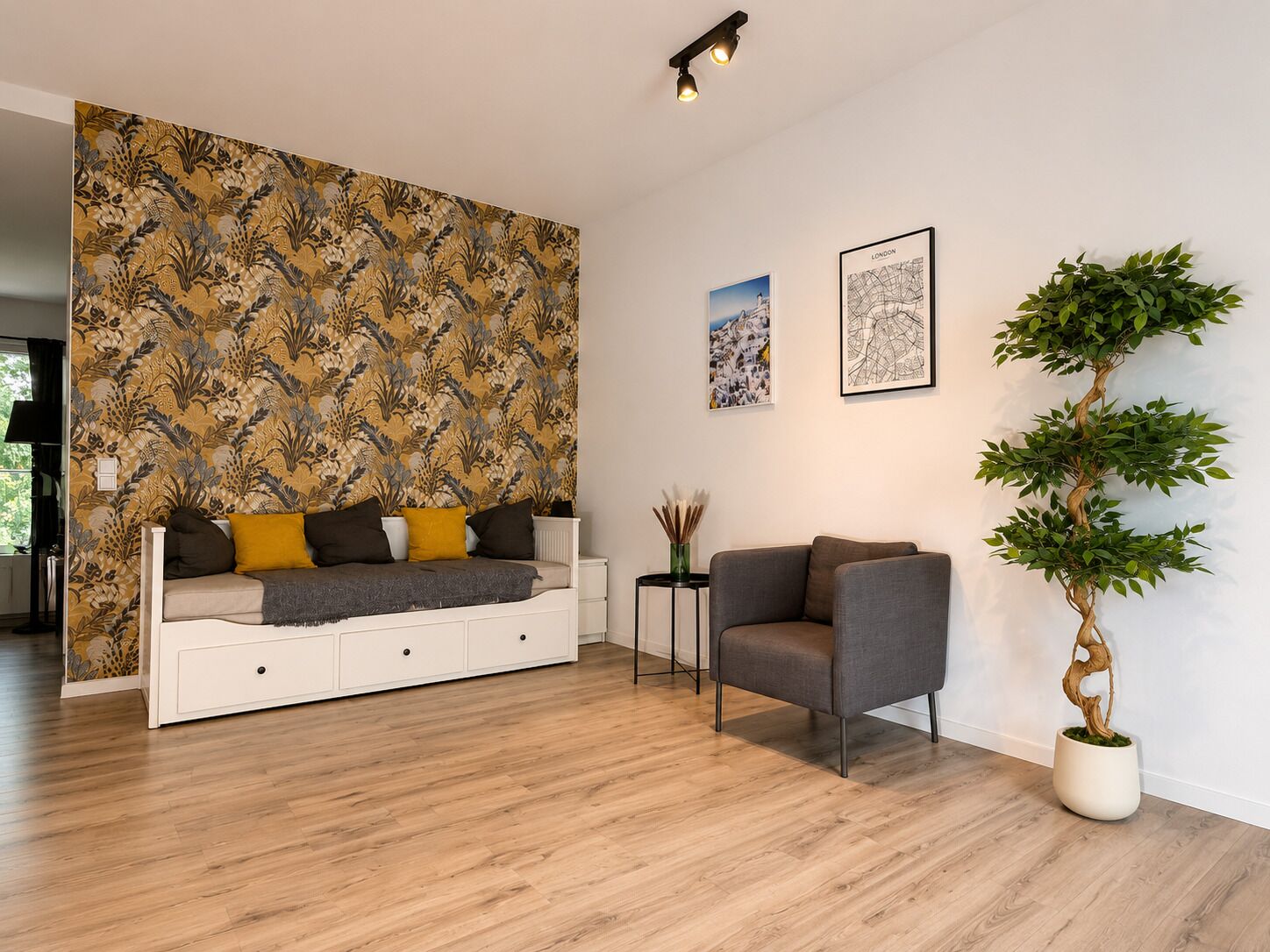 A modern living room with a patterned wallpaper, a sofa with cushions, a grey armchair, a potted plant, and two framed pictures on the wall.