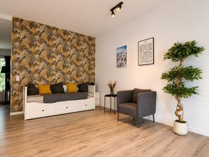 Una sala de estar moderna con papel tapiz estampado, un sofá con cojines, un sillón gris, una planta en maceta y dos cuadros enmarcados en la pared.