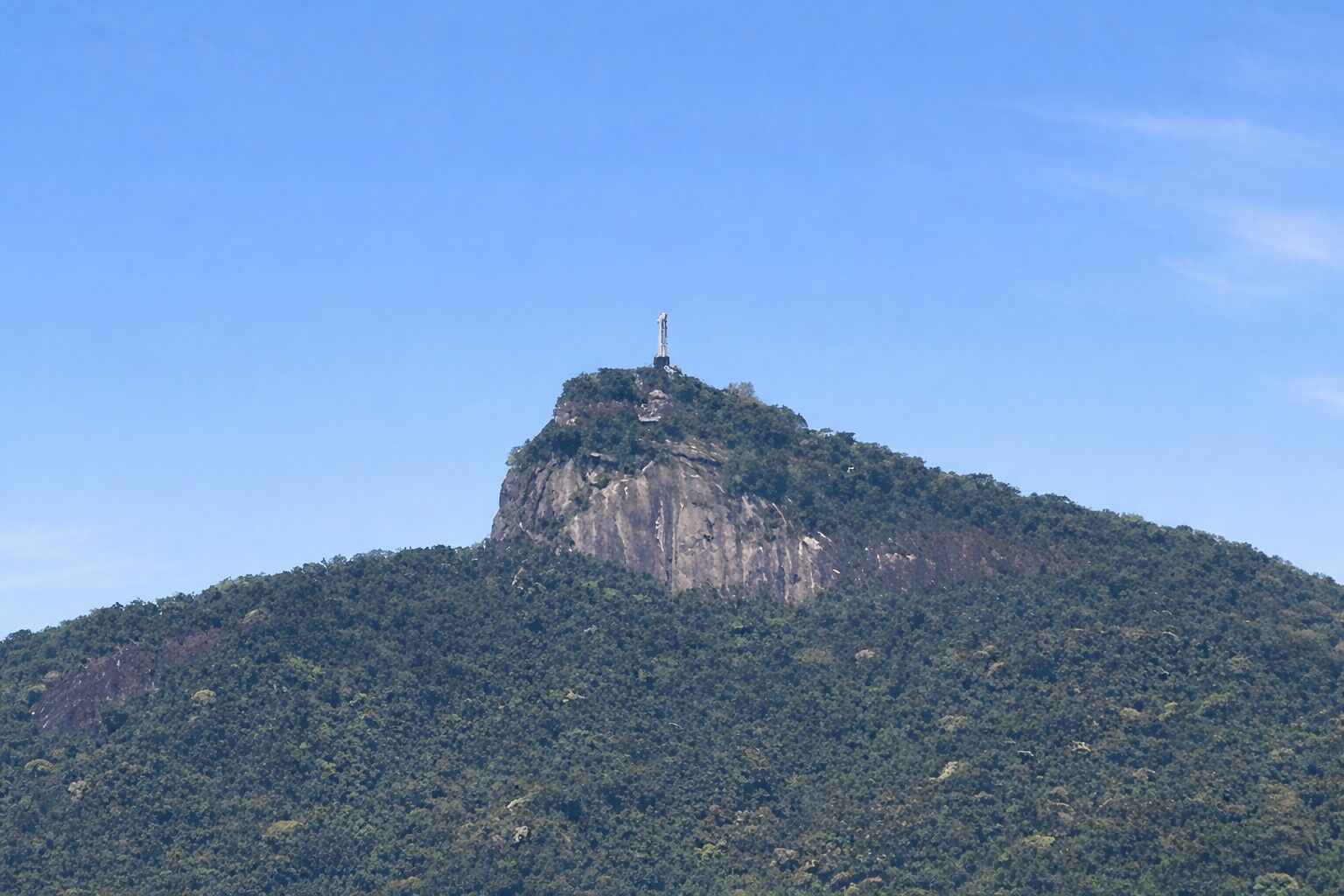 A mountain with a statue on top, surrounded by dense forest.