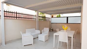 Una veranda esterna coperta con mobili in rattan bianco, tra cui un divano, sedie e un tavolo con un vaso di fiori.