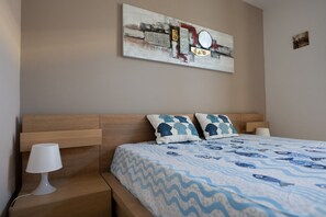 A bedroom with a wooden headboard, a bed with a blue and white patterned bedspread, two decorative pillows, a bedside table with a lamp, and a wall-mounted abstract painting. - Cozy 55 sqm apartment that can accommodate up to 4 guests, located on the ground floor of a building (accessible via a few steps). The property is situated in Pozzallo—a seaside town with beaches that have repeatedly been awarded the Blue Flag—in a q (Pozzallo)