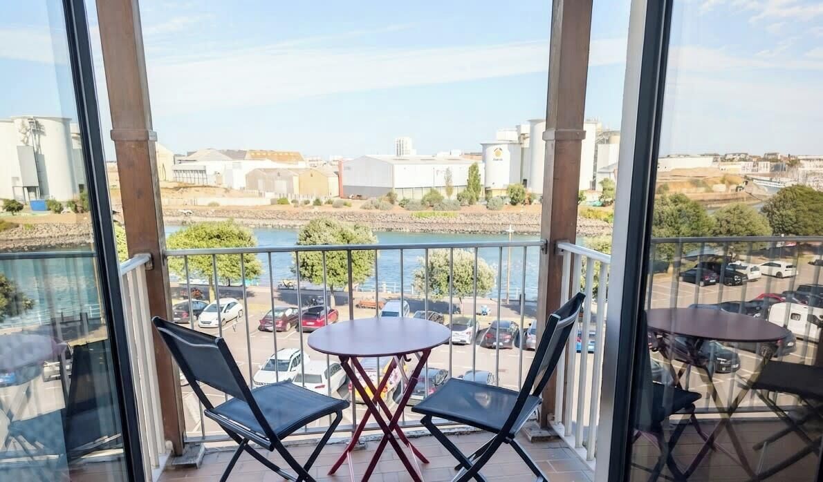 Un balcon avec une table et des chaises, donnant sur un parking et une rivière, avec des bâtiments en arrière-plan.