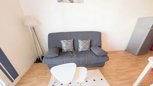 Ein Wohnzimmer mit grauem Sofa, zwei gemusterten Kissen, einem weißen Sessel, einer Lampe und einem Kühlschrank.