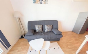 Ein Wohnzimmer mit grauem Sofa, zwei gemusterten Kissen, einem weißen Sessel, einer Lampe und einem Kühlschrank.