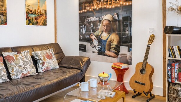 Un salon avec un canapé en cuir marron, une table basse en verre sur laquelle sont posés des magazines, une guitare et une bibliothèque.