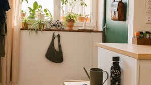 Une pièce avec une fenêtre, une étagère avec des plantes, une porte à laquelle est accroché un sac, une petite table avec une tasse, une bouteille d’eau et une paire de lunettes de soleil.