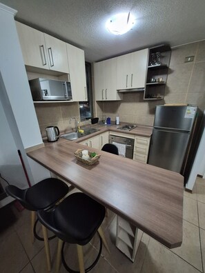 A compact kitchen with a wooden countertop, two black chairs, a stainless steel refrigerator, and a microwave above the sink. - Cozy Apartment with a Balcony, Pool, and Beautiful View in Valparaíso (Valparaíso)