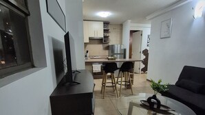 Ett modernt vardagsrum med ett soffbord i glas, en svart soffa och en tv på ett tv-bord. I bakgrunden finns ett köksområde med ett matbord och stolar.