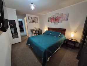 Una habitación de hotel con cama, escritorio, silla, espejo y obras de arte en las paredes.