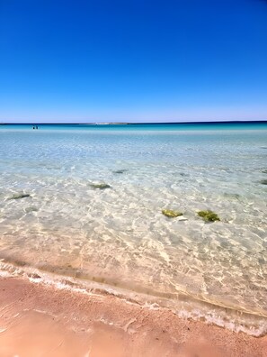 Una playa con aguas cristalinas y poco profundas, un muelle distante y cielo despejado.