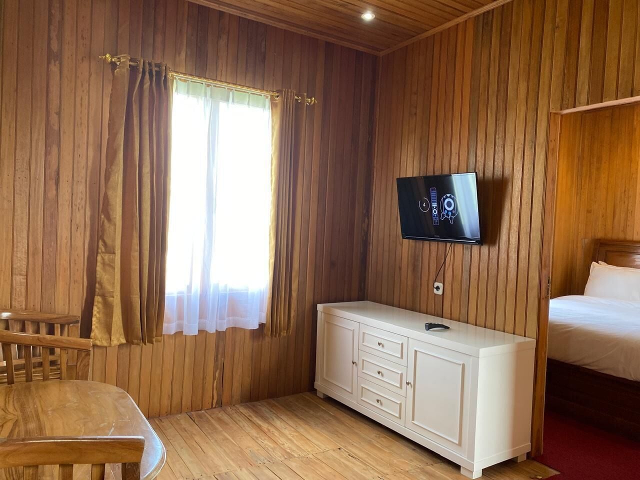 Huoneessa on puupaneloidut seinät, sänky, seinälle asennettu televisio, valkoinen lipasto ja puinen tuoli.