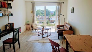Ein Wohnzimmer mit einer Couch, einem Esstisch, einem Couchtisch, einem Bücherregal, einem Sessel und Blick durch ein großes Fenster.