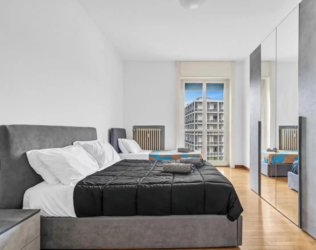 Ein modernes Schlafzimmer mit einem großen Bett, einem Nachttisch, einer Kommode und Blick auf ein Gebäude durch ein Fenster.