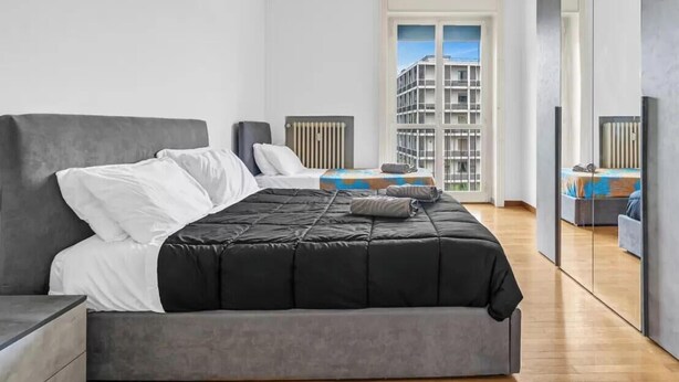 Una camera da letto moderna con un letto grande, un comodino, una cassettiera e vista su un edificio attraverso una finestra.